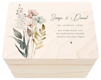 Könnte beinhalten: Eine Holzkiste mit einer hellen Holzoptik und einem floralen Design auf dem Deckel. Der Deckel trägt den Text "Svenja & Daniel" und "20. AUGUST 2026" in grauer Schrift. Der Text "Aus Liebe macht man die komischsten Sachen, manche heiraten sogar!" steht ebenfalls in grauer Schrift auf dem Deckel.