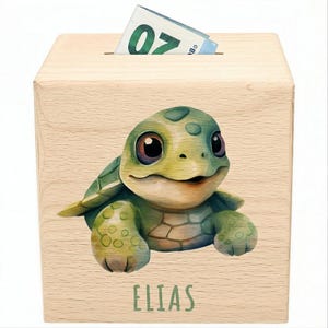 Könnte beinhalten: Eine Holzspardose mit einem Schlitz oben, mit einer Cartoon-Schildkröten-Illustration. Die Schildkröte ist grün und hellbraun mit großen Augen. Der Name "ELIAS" ist unten grün aufgedruckt. Ein 20-Euro-Schein ist teilweise sichtbar.
