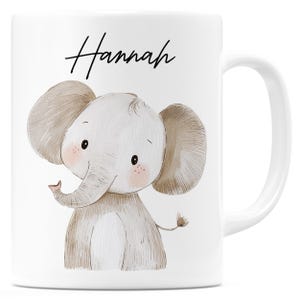 Könnte beinhalten: Weiße Keramiktasse mit einer Illustration eines Cartoon-Elefanten. Der Elefant ist grau und hat große Ohren und einen Rüssel. Der Name "Hannah" steht in schwarzer Schrift über dem Elefanten. Die Tasse hat einen gebogenen Griff.