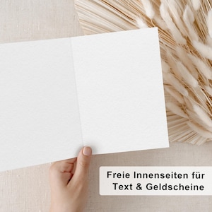 May include: A blank white card with a textured surface. The card is folded in half and is being held by a hand. The text "Freie Innenseiten f&uuml;r Text & Geldgeschenke" is visible at the bottom of the image.