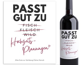 May include: A white card with black text that reads "PASST GUT ZU" with lines drawn through "FISCH" and "FLEISCH" and "WILD" written below. The text "Hochzeits-Planungen" is written in pink below the lines. The card also has the text "Alles Gute zur Verlobung! Deine Hannah" at the bottom. A bottle of red wine with a white label that reads "PASST GUT ZU" with lines drawn through "FISCH" and "FLEISCH" and "WILD" written below. The text "Hochzeits-Planungen" is written in pink below the lines. The label also has the text "Alles Gute zur Verlobung! Deine Hannah" at the bottom.