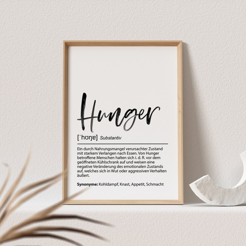 May include: A framed print with the word "Hunger" in a handwritten style. Below the word is a definition in German. The frame is a light wood color, and the background is white.