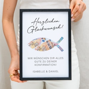 May include: A black framed picture with a white background. The picture has a cutout of a fish with money inside. The text on the picture reads "Herzlichen Gl&uuml;ckwunsch!" and "Wir w&uuml;nschen dir alles gute zu deiner Konfirmation! Isabelle & Daniel".