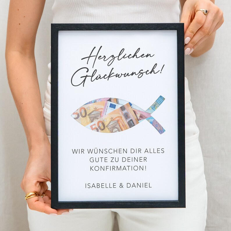May include: A black framed picture with a white background. The picture has a cutout of a fish with money inside. The text on the picture reads "Herzlichen Gl&uuml;ckwunsch!" and "Wir w&uuml;nschen dir alles gute zu deiner Konfirmation! Isabelle & Daniel".