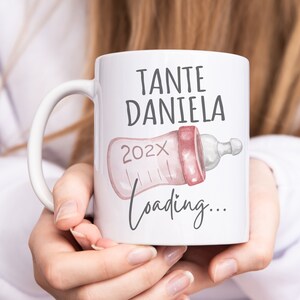 Könnte beinhalten: Weiße Keramiktasse mit einem rosa und grauen Babyflaschen-Design. Der Text auf der Tasse lautet "Tante Daniela 202X Loading..."