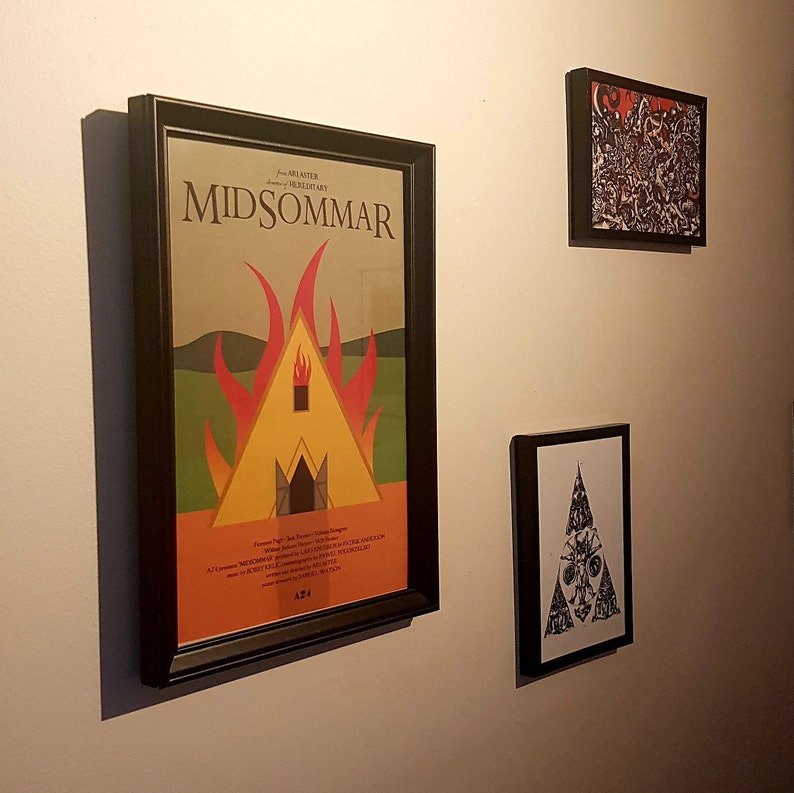 May include: Three framed movie posters on a white wall. The largest poster is black with a yellow and red image of a burning house. The poster has the text "MIDSUMMER" in large black letters. The other two posters are smaller and have black and white images.
