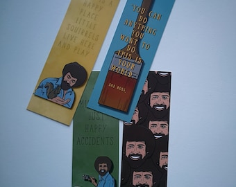 May include: Set of three bookmarks featuring Bob Ross, a famous painter. The bookmarks are printed with quotes from Bob Ross and images of him. The first bookmark is yellow and features a quote about a happy place. The second bookmark is blue and features a quote about doing anything you want to do. The third bookmark is green and features a quote about happy accidents. The bookmarks are all rectangular and have rounded corners.