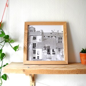 May include: A black and white line drawing of a European city scene with multiple buildings, rooftops, and windows. The drawing is framed in a light wood frame and is displayed on a wooden shelf.