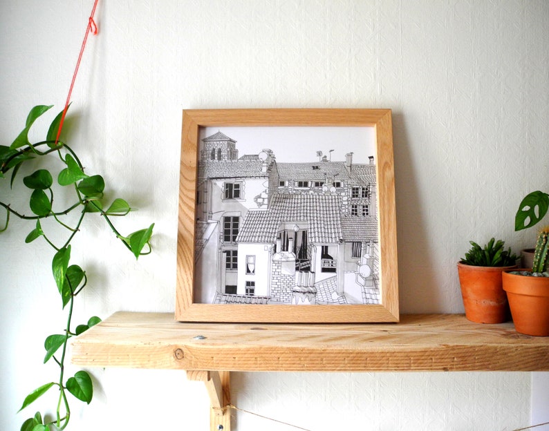 May include: A black and white line drawing of a European city scene with multiple buildings, rooftops, and windows. The drawing is framed in a light wood frame and is displayed on a wooden shelf.