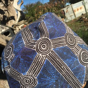 May include: A blue and black patterned head covering. The design features white dotted lines and circular patterns. The background is blurred, with trees and a blue sky visible.