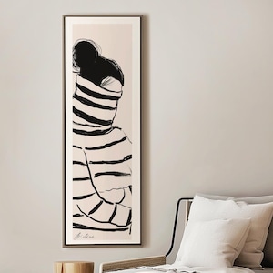 Puede incluir: Un dibujo en blanco y negro de una persona con un suéter a rayas, enmarcado en un marco de madera clara. El marco está colgado en una pared blanca sobre un taburete de madera y un sofá blanco con una manta blanca.