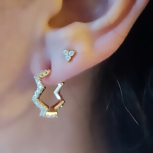 May include: A pair of gold earrings with a zig-zag design and clear cubic zirconia stones. The earrings are shown in a person's ear.