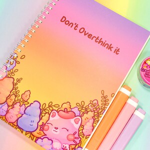May include: A pink and orange gradient notebook with the text "Don't Overthink it" in red. The notebook has a spiral binding and a cute cartoon cat illustration on the bottom left corner.