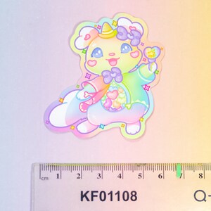 以下が含まれることがあります： 帽子をかぶって杖を持った、パステルレインボーカラーの漫画の動物のステッカー。動物はピンクのリボンとハート型の鼻をしています。ステッカーの幅は6cmです。