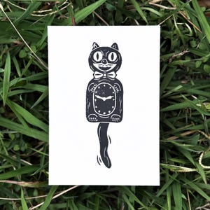 May include: A black and white cat-shaped clock design on a white card. The clock features a bow tie, round eyes, and a pendulum tail. The card is set against a backdrop of green grass.