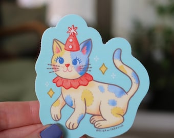 May include: A die-cut sticker on a light blue background featuring a cream-colored cat with blue and yellow patches. The cat wears a red clown collar and a matching red party hat with white dots. Small yellow stars surround the cat illustration.