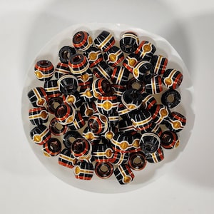 Peut inclure: Une collection de petites perles en forme de tonneau avec une base noire brillante et des rayures horizontales rouges, blanches et dor&eacute;es. Chaque perle pr&eacute;sente un trou central et un motif r&eacute;p&eacute;titif de cercles dor&eacute;s sur la section m&eacute;diane.
