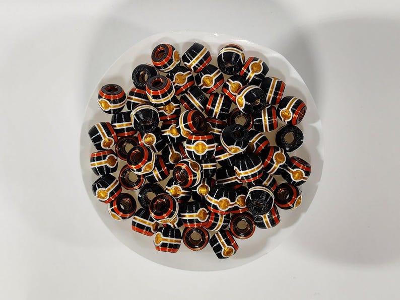 Peut inclure: Une collection de petites perles en forme de tonneau avec une base noire brillante et des rayures horizontales rouges, blanches et dor&eacute;es. Chaque perle pr&eacute;sente un trou central et un motif r&eacute;p&eacute;titif de cercles dor&eacute;s sur la section m&eacute;diane.