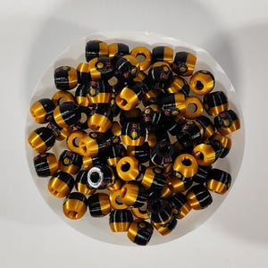 Peut inclure: Un ensemble de petites perles en forme de tonneau pr&eacute;sentant un design bicolore avec des sections en or m&eacute;tallique et en noir brillant. Chaque perle poss&egrave;de un trou central et une petite lettre R rouge imprim&eacute;e sur la partie noire.