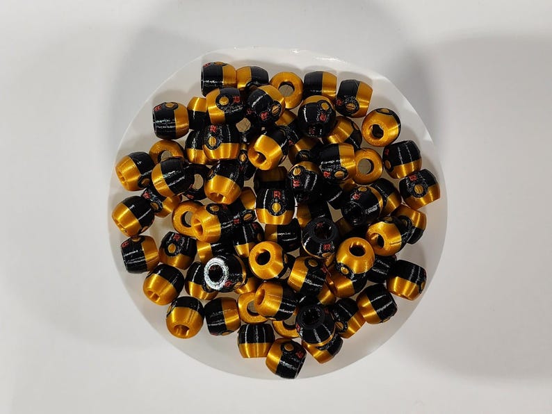 Peut inclure: Un ensemble de petites perles en forme de tonneau pr&eacute;sentant un design bicolore avec des sections en or m&eacute;tallique et en noir brillant. Chaque perle poss&egrave;de un trou central et une petite lettre R rouge imprim&eacute;e sur la partie noire.