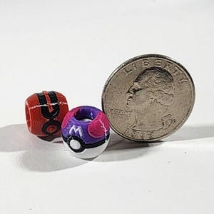 Peut inclure: Deux petites perles sph&eacute;riques con&ccedil;ues pour ressembler &agrave; des r&eacute;cipients miniatures en forme de boule. Une perle est rouge et noire avec une bande centrale, l'autre est violette, rose et blanche avec une lettre M. Une pi&egrave;ce est visible pour l'&eacute;chelle.