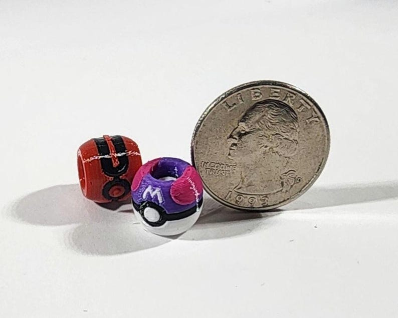 Peut inclure: Deux petites perles sph&eacute;riques con&ccedil;ues pour ressembler &agrave; des r&eacute;cipients miniatures en forme de boule. Une perle est rouge et noire avec une bande centrale, l'autre est violette, rose et blanche avec une lettre M. Une pi&egrave;ce est visible pour l'&eacute;chelle.