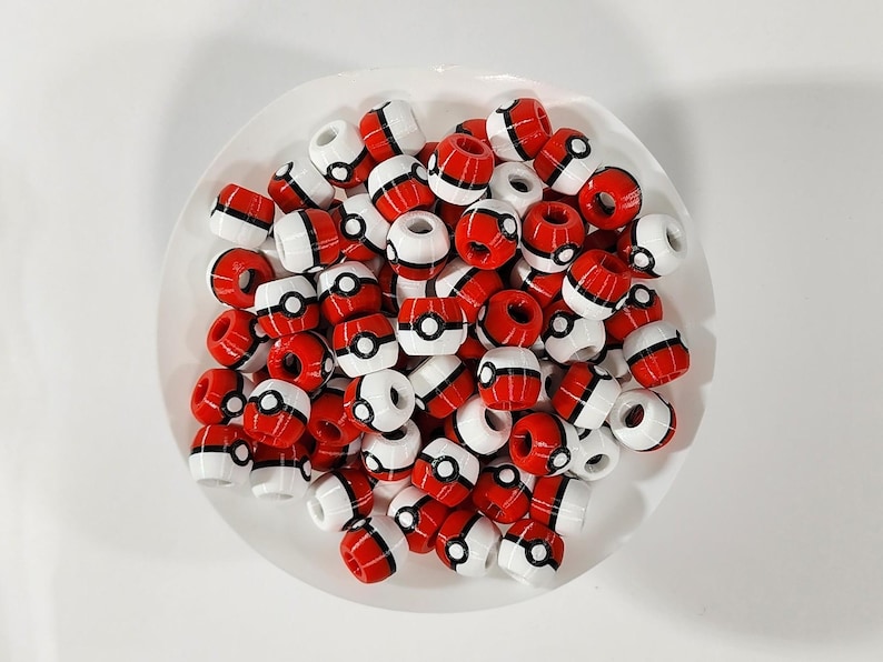 Peut inclure: Une collection de petites perles sph&eacute;riques pr&eacute;sentant un motif rouge et blanc avec une bande horizontale noire et un d&eacute;tail circulaire central noir et blanc, rappelant un design de balle, empil&eacute;es dans un r&eacute;cipient blanc circulaire.