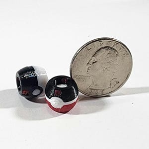 Peut inclure: Deux petites perles sph&eacute;riques con&ccedil;ues pour ressembler &agrave; des Pok&eacute; Balls, l'une noire et blanche et l'autre noire, blanche et rouge. Une pi&egrave;ce de monnaie am&eacute;ricaine de 25 cents est plac&eacute;e &agrave; c&ocirc;t&eacute; des perles pour montrer leur petite taille.