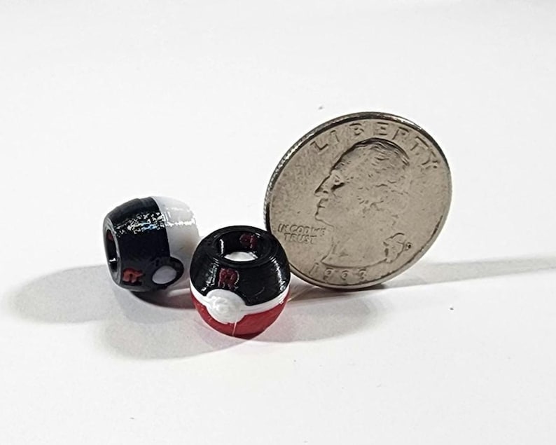 Peut inclure: Deux petites perles sph&eacute;riques con&ccedil;ues pour ressembler &agrave; des Pok&eacute; Balls, l'une noire et blanche et l'autre noire, blanche et rouge. Une pi&egrave;ce de monnaie am&eacute;ricaine de 25 cents est plac&eacute;e &agrave; c&ocirc;t&eacute; des perles pour montrer leur petite taille.