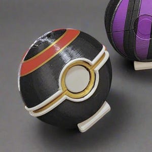 K&ouml;nnte beinhalten: Ein dekorativer schwarz-goldener Ball mit einem wei&szlig;en kreisf&ouml;rmigen Mittelteil und einem roten Streifen. Im Hintergrund befindet sich ein zweiter Ball mit lila und schwarzen Streifen. Die B&auml;lle liegen auf einer grauen Oberfl&auml;che.
