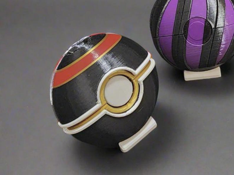 K&ouml;nnte beinhalten: Ein dekorativer schwarz-goldener Ball mit einem wei&szlig;en kreisf&ouml;rmigen Mittelteil und einem roten Streifen. Im Hintergrund befindet sich ein zweiter Ball mit lila und schwarzen Streifen. Die B&auml;lle liegen auf einer grauen Oberfl&auml;che.