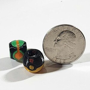 Peut inclure: Deux petites perles sph&eacute;riques aux motifs d&eacute;taill&eacute;s. Une perle est noire avec des accents verts et orange, tandis que l'autre est noire et dor&eacute;e avec une lettre R rouge. Une pi&egrave;ce de 25 cents est plac&eacute;e &agrave; c&ocirc;t&eacute; pour montrer leur petite taille.