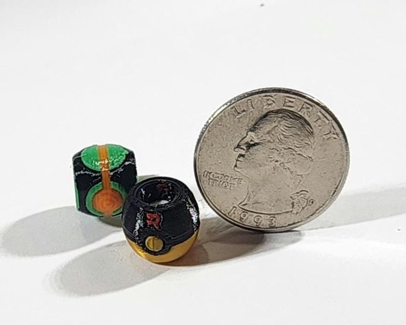 Peut inclure: Deux petites perles sph&eacute;riques aux motifs d&eacute;taill&eacute;s. Une perle est noire avec des accents verts et orange, tandis que l'autre est noire et dor&eacute;e avec une lettre R rouge. Une pi&egrave;ce de 25 cents est plac&eacute;e &agrave; c&ocirc;t&eacute; pour montrer leur petite taille.