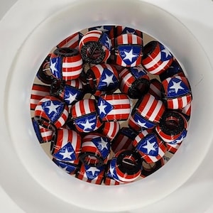 May include: A collection of small, cylindrical beads with a patriotic design. The beads feature alternating red and white stripes, blue sections with a white star, and a dark interior. They are contained within a white, circular container.