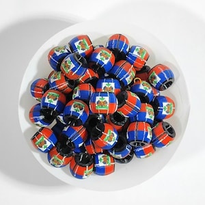 以下が含まれることがあります： 白いボウルに入った樽型のビーズのコレクション。ビーズは主に赤、青、白で、紋章のデザインが施されています。ビーズはおそらく、ジュエリー作りや工芸品に使われます。