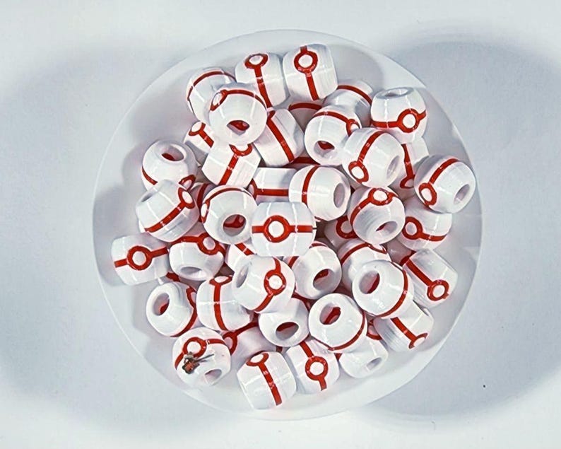 Peut inclure: Une collection de perles blanches en forme de tonneau, orn&eacute;es d'une bande rouge et d'un cercle rouge central, rappelant un r&eacute;cipient sph&eacute;rique. Ces petites perles cylindriques en plastique sont empil&eacute;es, chacune perc&eacute;e en son centre.