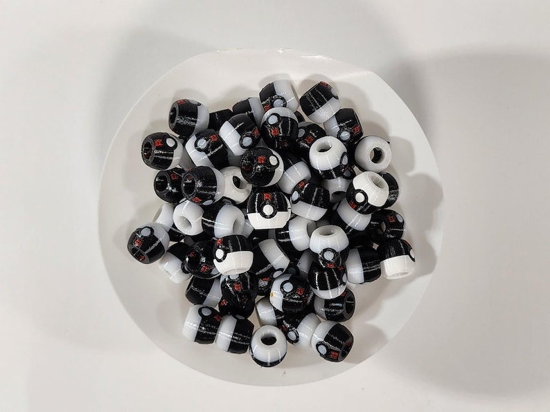 Peut inclure: Une collection de petites perles en plastique en forme de tonneau avec un motif noir et blanc. Chaque perle pr&eacute;sente une ligne horizontale centrale, un petit d&eacute;tail circulaire et une touche rouge, rappelant un contenant sph&eacute;rique.