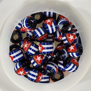 以下が含まれることがあります： キューバの国旗にインスパイアされたデザインの円筒形ビーズのコレクション。ビーズは、青と白のストライプが交互に配置され、白い星が表示された赤い部分があります。ビーズは白いボウルの中にあります。