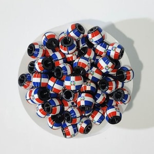 以下が含まれることがあります： 赤、白、青のストライプと小さなエンブレムが特徴の円筒形のビーズのコレクション。ビーズは白いボウルに配置されており、視覚的に魅力的なディスプレイを作成しています。