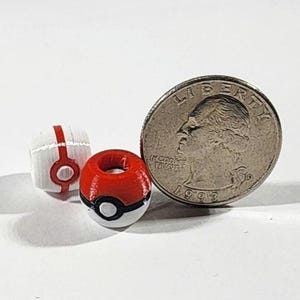 Peut inclure: Deux petites perles sph&eacute;riques con&ccedil;ues pour ressembler &agrave; des Pok&eacute; Balls rouges, blanches et noires, plac&eacute;es &agrave; c&ocirc;t&eacute; d'une pi&egrave;ce de 25 cents am&eacute;ricaine pour l'&eacute;chelle. Les perles pr&eacute;sentent un trou central et un motif circulaire imprim&eacute;.