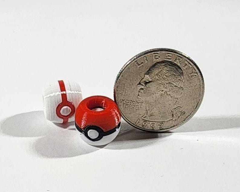 Peut inclure: Deux petites perles sph&eacute;riques con&ccedil;ues pour ressembler &agrave; des Pok&eacute; Balls rouges, blanches et noires, plac&eacute;es &agrave; c&ocirc;t&eacute; d'une pi&egrave;ce de 25 cents am&eacute;ricaine pour l'&eacute;chelle. Les perles pr&eacute;sentent un trou central et un motif circulaire imprim&eacute;.