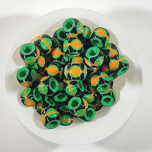 Peut inclure: Une collection de petites perles cylindriques pr&eacute;sentant un ext&eacute;rieur noir avec des accents circulaires vert vif et orange. Chaque perle poss&egrave;de un trou central et une surface textur&eacute;e, dispos&eacute;es en groupe pour montrer le contraste.