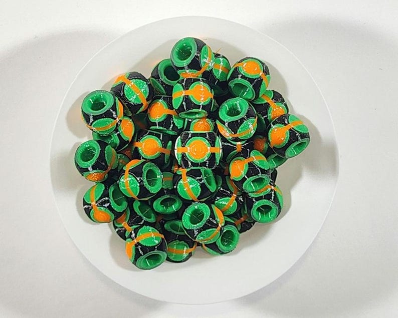 Peut inclure: Une collection de petites perles cylindriques pr&eacute;sentant un ext&eacute;rieur noir avec des accents circulaires vert vif et orange. Chaque perle poss&egrave;de un trou central et une surface textur&eacute;e, dispos&eacute;es en groupe pour montrer le contraste.