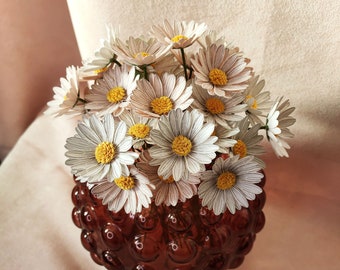 Könnte beinhalten: Ein Strauß weißer Papiermargeriten mit gelben Zentren, die in einer braunen, blasenförmigen Vase angeordnet sind.