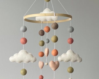 Peut inclure: Mobile pour bébé avec un anneau en bois et des formes en feutre suspendues. Le mobile présente des nuages blancs, des sphères roses et oranges, et des formes de cœur. La palette de couleurs comprend des nuances de rose, d'orange, de vert et de gris.