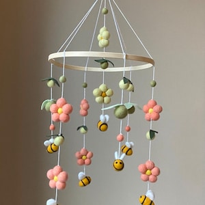 May include: A wooden baby mobile with felt flowers, bees, and leaves. The flowers are pink and yellow, and the bees are yellow with black stripes. The mobile is hanging from a wooden ring.
