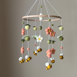 May include: A baby mobile featuring a wooden ring with hanging felt elements. The mobile includes green apples, pink flowers, white daisies, and yellow and black bees. The mobile is suspended by white strings.