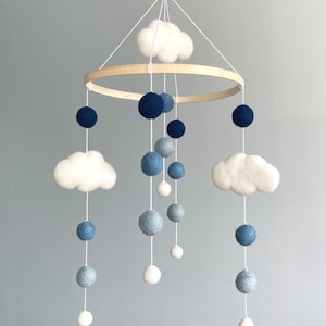 May include: A hanging mobile featuring a circular wooden frame with white felt clouds and felt balls in shades of navy blue, light blue, grey, and white. The elements are suspended by white strings, creating a layered arrangement of soft textures.