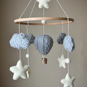 May include: A hanging mobile featuring a central grey hot air balloon, surrounded by textured grey clouds and off-white felt stars suspended from a circular wooden frame. White strings connect the elements, with small white felt balls as accents.