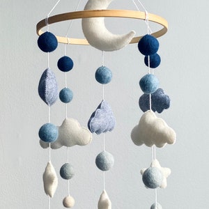 May include: A white felt crescent moon hangs from a wooden ring with blue felt balls and clouds. The mobile also features white and blue felt stars and raindrops.